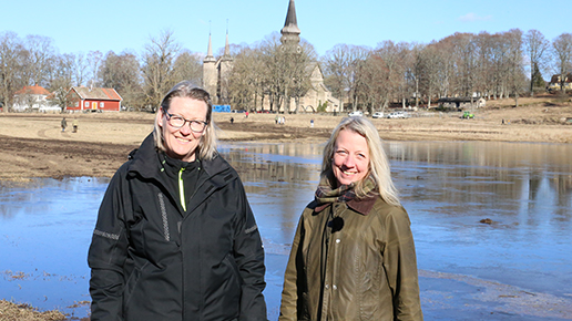 Maria Vretemark och Marie Wennerholm står framför den nybildade våtmarken med kulturmiljön runt klostret i bakgrunden.