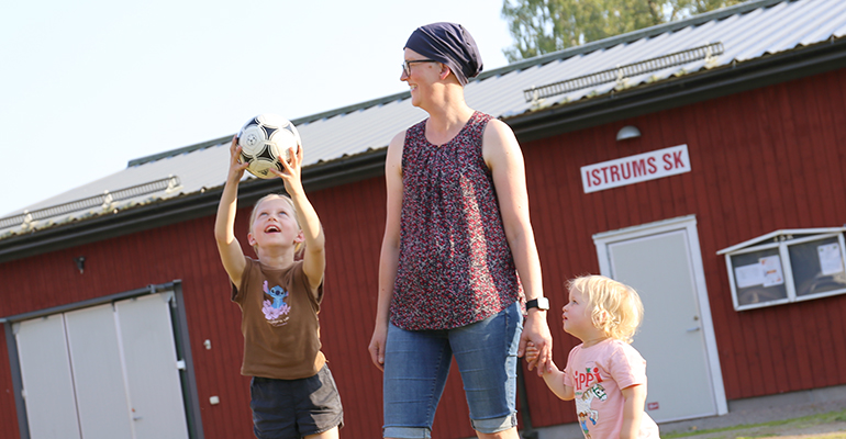 En mamma med två barn framför en föreningslokal. Den äldre dottern fångar en boll.