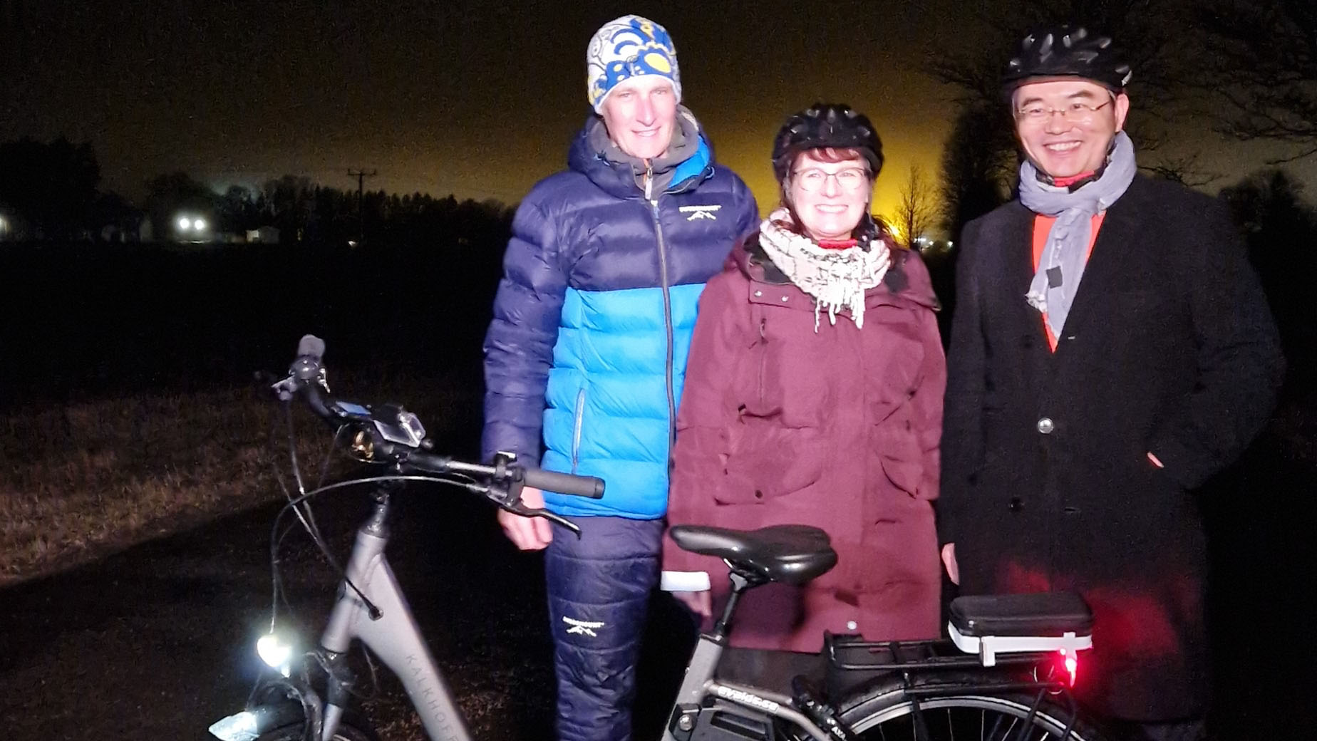 Tre glada personer står bakom en cykel. Det är mörkt ute