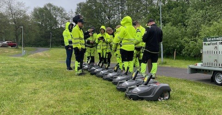 Nio robotgräsklippare på en gräsmatta. Ett tiotal personer med gula reflexkläder står runt omkring.