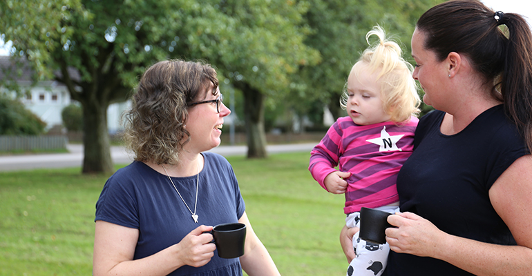 Två kvinnor dricker kaffe vid en lekplats. Ett litet barn sitter på sin mammas arm.