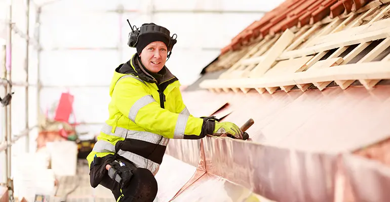 En person i gula arbetskläder står vid ett tak som ser ut att byggas. Röda takpannor syns längst upp i bild.
