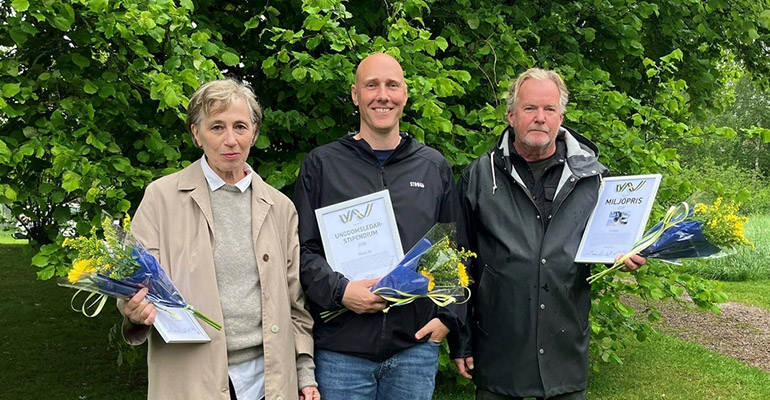 Tre personer står framför en träd med gröna blad. De tittar in i kameran och har diplom och blombuketter i händerna. 