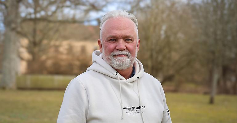 Porträtt av en man med grått skägg klädd i en ljus hoodie, fotograferad utomhus.
