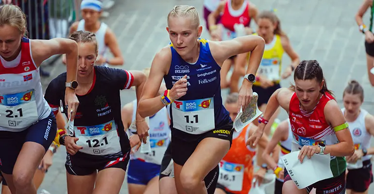 Kvinnliga orienterare springer och ser koncentrerade ut. I mitten springer en person i blont hår och det står "Sweden" på hennes kläder.