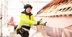 En person i gula arbetskläder står vid ett tak som ser ut att byggas. Röda takpannor syns längst upp i bild.