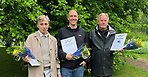 Tre personer står framför en träd med gröna blad. De tittar in i kameran och har diplom och blombuketter i händerna. 