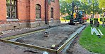Brädor som ramar in påbörjad boulebana. En grävmaskin står längre bak i bild. En röd tegelvägg på stadshuset syns till vänster.