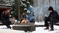 Fyra ungdomar sitter vid en grillplats. En eld är tänd och det är fullt med snö på marken och granarnas grenar.